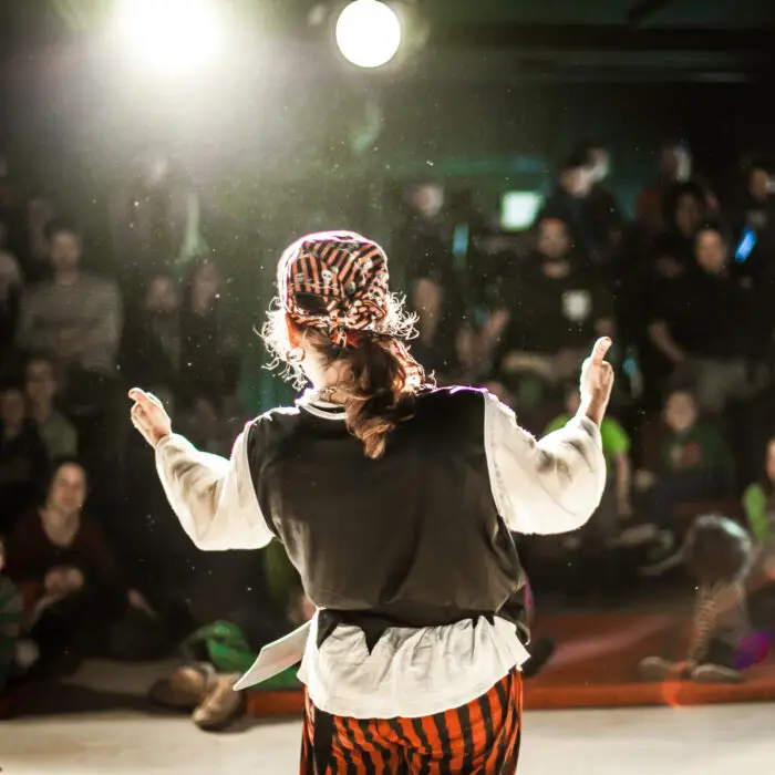 A performing arts entertainer is seen from the back in selective focus, dressed as a pirate on stage during a comedy act with blurry audience at back.